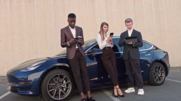 Tres jóvenes de éxito en el fondo de un coche eléctrico deportivo, el uso de aparatos de teléfono y tableta. la tecnología y la gente concept.Business gente. Prosperidad. — Vídeos de Stock