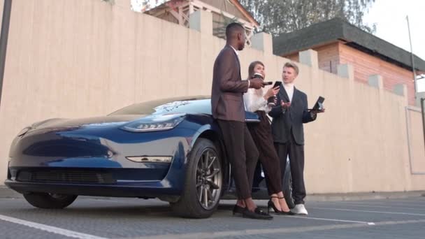 Vendedor presentando un coche nuevo a una joven pareja al aire libre. El distribuidor acertado del coche le dice a los dueños sobre los beneficios de su coche en estacionamiento cerca de concesionario del coche. Movimiento lento — Vídeos de Stock