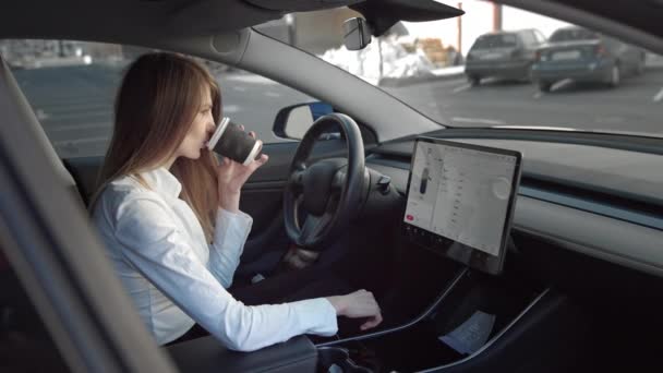 Портрет молодої красивої успішної бізнес-леді, яка сидить у дорогому спортивному електричному автомобілі, п'є каву перед офісом, використовує сенсорний екран автомобіля для пошуку інформації — стокове відео