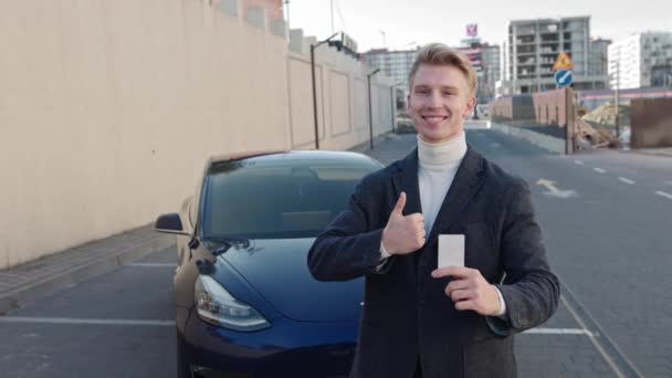 Ближе к делу уверенный молодой блондин покупает новую машину. Веселый бизнесмен держит в руках современный электронный ключ от машины, показывает жест ОК, замедленная съемка. Портрет успешного человека, движущегося к своей цели — стоковое видео