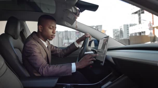 Yan görüş. Genç, siyah bir işadamı elinde kahveyle modern bir arabada oturuyor. Bakışlarını kameraya çeviriyor. Elektrikli arabada başarılı bir iş adamı. — Stok video