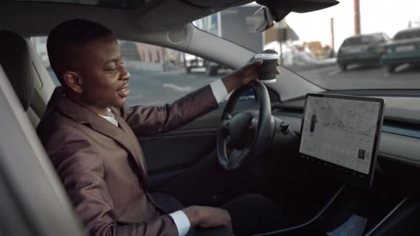 Stilren ung afrikansk amerikan med kaffe i händerna, sitter i en modern sport elbil tittar på skärmen, tittar på kameran. Sömnig affärsman, sidovy. — Stockvideo