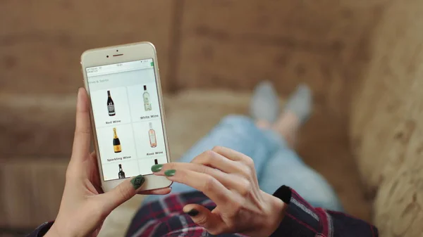 Femme à la maison Allongé sur le canapé dans le salon en utilisant Smartphone achète dans Internet Shop. Femme commande des vins à la maison dans une boutique en ligne à l'aide d'un smartphone. Female Selects Wines in Grocery Online Store. Point de vue Images De Stock Libres De Droits