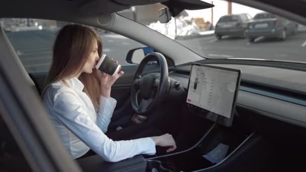 Retrato de uma jovem bela empresária bem sucedida sentada em um carro elétrico caro esportes, beber café em frente ao escritório, usa a tela sensível ao toque do carro para procurar informações — Vídeo de Stock