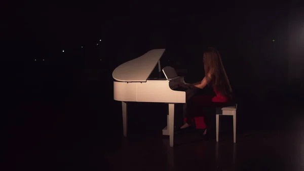 Uma jovem menina bonita em um vestido vermelho se aproxima do piano branco e se senta. Connet de música clássica. Salão atmosfera romântica cheio de fumaça — Fotografia de Stock