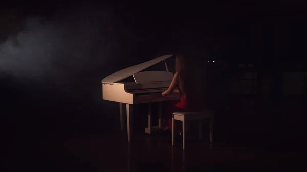 На концерте пианист играет на красивом белом рояле. Вид сзади. Молодая красивая девушка в красном платье и играет на пианино в черной комнате с легким ощущением дыма — стоковое фото