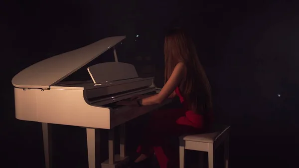 Pianista suona bellissimo pianoforte a coda bianco sul palco in concerto. Vista dal back.Young bella ragazza in abito rosso e suona il pianoforte in una stanza nera con una leggera sensazione di fumo — Foto Stock