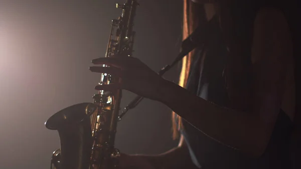Joven chica hermosa en un vestido oscuro juega en un saxofón brillante dorado en el escenario. Estudio oscuro con humo e iluminación de escenario. Las manos y el saxofón de cerca. Vista lateral —  Fotos de Stock