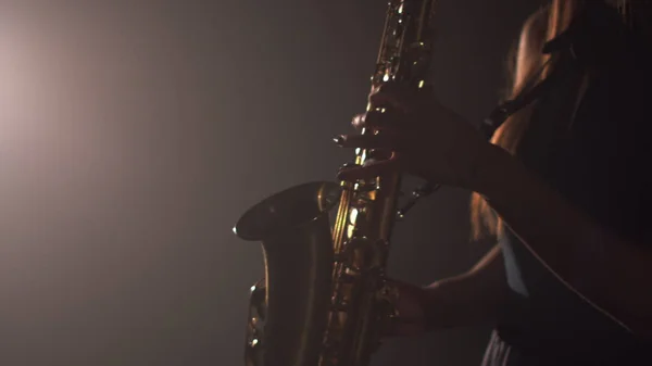 Jeune belle fille dans une robe sombre joue sur un saxophone doré brillant sur scène. Studio sombre avec fumée et éclairage scénique. Les mains et le saxophone se rapprochent. Vue latérale — Photo