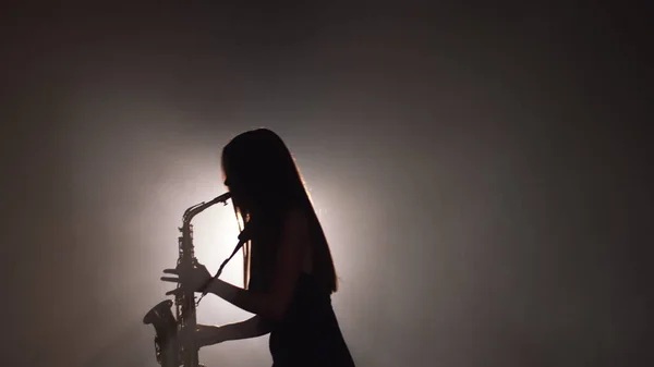 Young beautiful girl in a dark dress plays on a golden shiny saxophone on stage. Dark studio with smoke and stage lighting. Hands and saxophone close up. Side view.Slow motion video — Stock Photo, Image