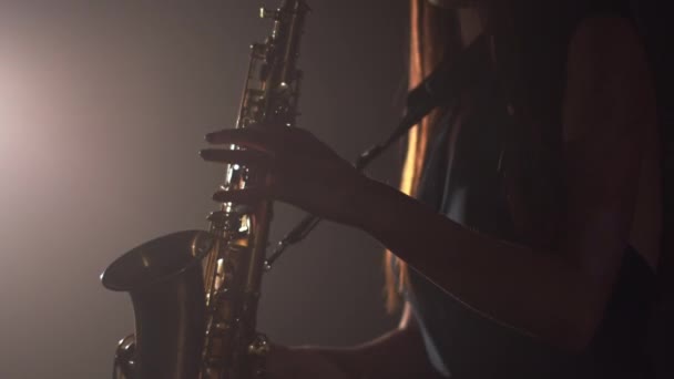 Молода красива дівчина в темній сукні грає на золотому блискучому саксофоні на сцені. Темна студія з димом і освітленням сцени. Руки і саксофон крупним планом. Вид збоку — стокове відео