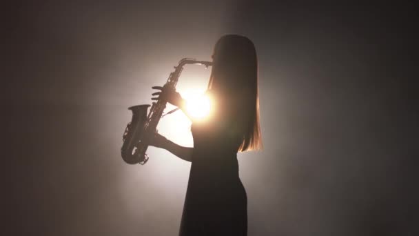 Young beautiful girl in a dark dress plays on a golden shiny saxophone on stage. Dark studio with smoke and stage lighting. Hands and saxophone close up. Side view.Slow motion video — Stock Video