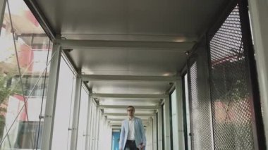 Şık ceketli ve gözlüklü genç bir adam modern cam bir koridorda kameraya gider. Kendine güvenen işadamı başarı toplantısına gider. iş, zafer konsepti 4k