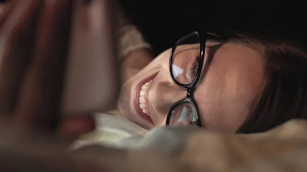 Donna con gli occhiali guarda smartphone e si trova sul letto, sorride sul viso — Foto Stock