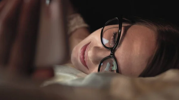 Mujer con gafas mira el teléfono inteligente y se encuentra en la cama, sonrisa en la cara — Foto de Stock