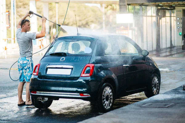 En ung, kjekk mann vasker bilen i bilvasken. Sommerbilvask. Bilvask under reise stockbilde