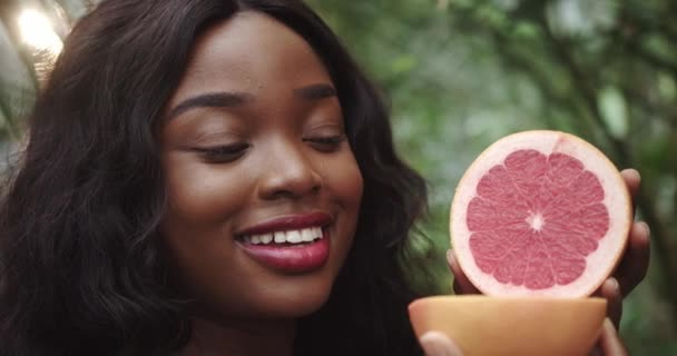 Foto de una encantadora joven africana sosteniendo rodajas de pomelo frente a su cara, posando frente a la cámara en una selva tropical. El concepto de cuidado saludable y de la piel. Movimiento lento — Vídeos de Stock