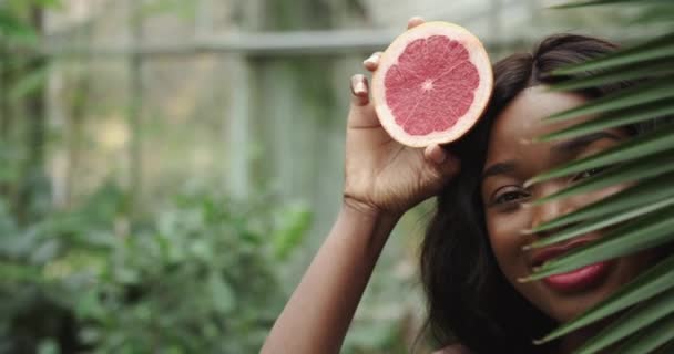 Primer plano de la cara y los ojos de la joven mujer afroamericana hermosa, piel oscura. Sostiene la mitad del pomelo cerca de su cara mirando a la cámara. Retrato de mujer de belleza natural saludable con fruta — Vídeos de Stock