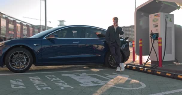 Hombre de negocios caucásico usando un teléfono inteligente y esperando a que la fuente de alimentación se conecte a los coches eléctricos para cargar la batería en el coche. Conecte el cargador del coche eléctrico de la carga — Vídeos de Stock