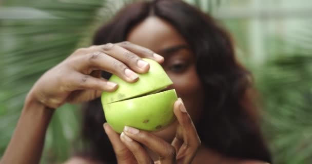 Ritratto di una giovane bella ragazza afroamericana che tiene tra le mani due metà di una mela davanti alla telecamera e le separa. Stile di vita sano, vitamine naturali per la cura del corpo. Girare dentro — Video Stock