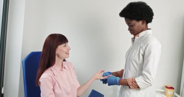 Análisis de sangre de dedos. Enfermera joven de apariencia afroamericana, toma un análisis de sangre de una chica de nacionalidad caucásica. Enfermera sacando sangre de una mano de mujer. Prueba médica del VIH, investigación — Vídeo de stock