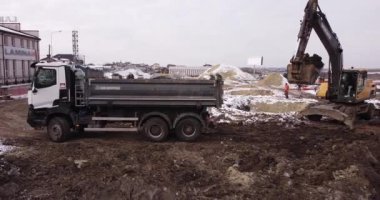 Üst hava çekimi: bir traktör kovası kamyon gövdesini doldurur. Bir kazıcı ve bir kamyon zemini dolduruyor. İşçilerin temel attığı inşaat alanının üzerinden uçuyor. Bir sürü ekipman