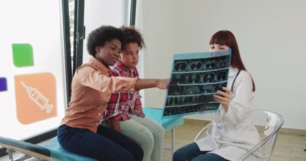 Baby mit der Mutter eines afroamerikanischen Mischlings auf einem Krankenhausempfang. Kinderärztin untersucht Kinder auf Röntgenbild mit schwarzer Mutter Schulmädchen Auf Ein Medical Visit. Gesundheitskonzept für Kinder — Stockvideo