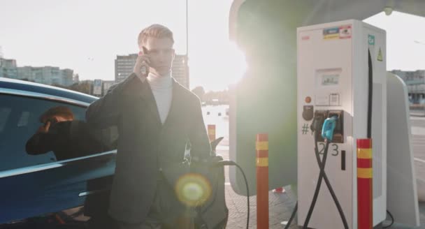 Empresario está hablando en su teléfono inteligente, mientras que su coche eléctrico está cargando en segundo plano. Vista lateral del coche de lujo azul. Vehículo eléctrico de carga masculina ambientalmente consciente. Estación de carga de coche — Vídeos de Stock