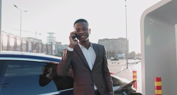 ビジネスマンアフリカ系アメリカ人は、彼の電気自動車が背景に充電している間、彼のスマートフォンで話しています。ブルー高級車のサイドビュー。環境に配慮した男性。充電ステーション — ストック動画