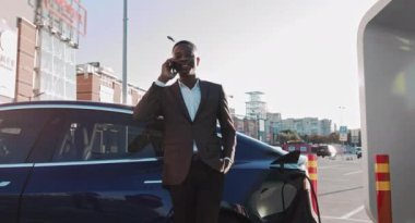 Özel donanımlı şarj istasyonunda lüks siyah araba şarj bataryası kullanırken hoş mobil sohbeti olan yakışıklı Afro-Amerikalı bir adamın portresi. Takım Elbiseli İş Adamı
