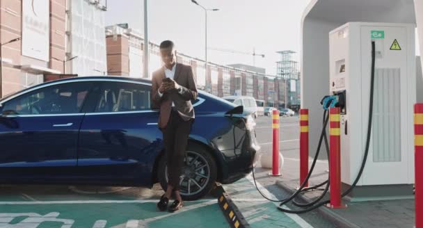 Homme d'affaires afro-américain utilisant un smartphone et s'appuie sur une voiture électrique de luxe blanche qui se charge. Prise de charge d'une voiture électrique. Les nouvelles technologies en ville s'arrêtent au centre-ville — Video