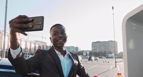 Αφροαμερικανός επιχειρηματίας χαμογελώντας νεαρός επιχειρηματίας Κάνοντας Selfie Video Call Μόνιμη Εξωτερική στο Business District. Πορτρέτο ενός blogger επιχειρηματίας στο σταθμό φόρτισης του ηλεκτρικού αυτοκινήτου — Αρχείο Βίντεο