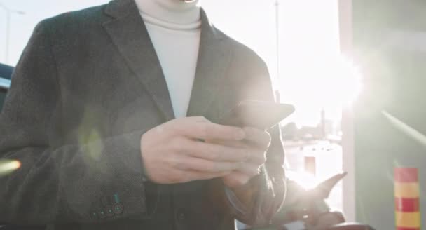 Kaukaski biznesmen w garniturze używa smartfona na ulicy w centrum miasta, zbliżenie telefonu. Mężczyzna stoi wokół elektrycznego samochodu na stacji ładowania. Przegląda Internet na swoim urządzeniu. — Wideo stockowe