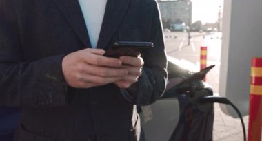 Kafkasyalı iş adamı akıllı telefon kullanıyor ve elektrikli araçlara aküyü şarj etmek için güç desteği bekliyor. Şarj istasyonundan bir elektrikli arabayı şarj etmek. Akıllı Telefon Ellerde