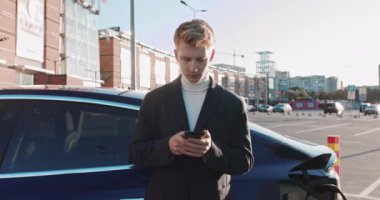 Elinde akıllı telefon olan bir işadamı, şarj olan mavi lüks elektrikli bir arabaya yaslanıyor. Elektrikli araba şarj prizi. Lüks elektrikli araba şarj oluyor. Adam Şarj İstasyonuna Bağlandı