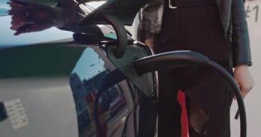 EV pili şarj etmek için elektrik kablosunu elektrikli bir arabaya bağlayan kadın. Elektrikli bir arabayı şarj ediyorum. Ağır çekim. Bir kadın eli şarj istasyonunun prizine dokunur.