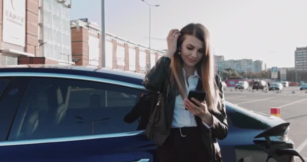 Avrupalı Kadın beklerken akıllı telefon kullanır ve pili şarj etmek için elektrikli araçlara güç kaynağı sağlar. Elektrikli araba şarj ediliyor.. — Stok video