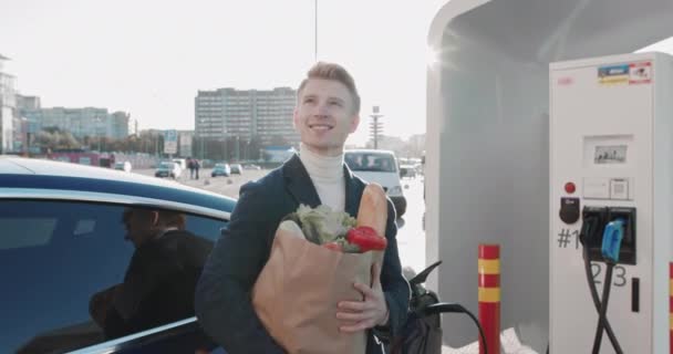 Flot blond fyr holder en madpakke nær en elektrisk bil, der oplades. Shopping i butikken. Elektrisk biloplader. Luksus elbil oplades. Pigen ser på kameraet og smiler – Stock-video