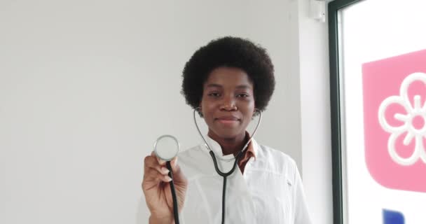 Close-up de mulher afro-americana Doutor em casaco branco na clínica no local de trabalho detém estetoscópio e aponta para a câmera. A doutora usa estetoscópio. Retrato de uma jovem médica. — Vídeo de Stock