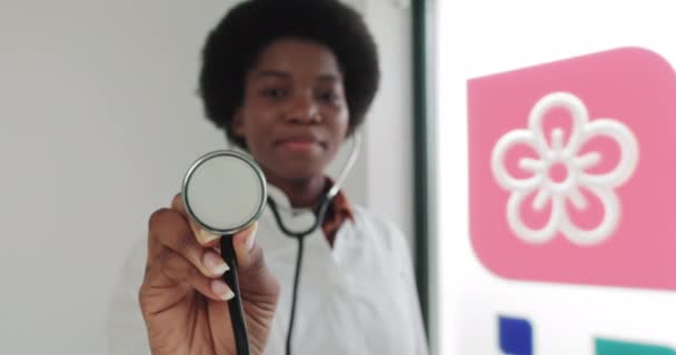 Η γυναίκα γιατρός χρησιμοποιεί στηθοσκόπιο. Κοντινό πλάνο της Αφροαμερικανίδας γιατρού με το λευκό παλτό στην κλινική στο χώρο εργασίας που κρατά στηθοσκόπιο. Το πορτρέτο μιας νεαρής γυναίκας γιατρού. Τόπος για το λογότυπο — Αρχείο Βίντεο