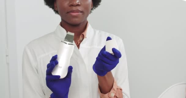 Portret van een vrouwelijke Afro-Amerikaanse cosmetoloog die een ultrasone gezichtsmachine in haar handen houdt tijdens de reinigingsprocedure. Niet-chirurgische facelift. dermatoloog — Stockvideo