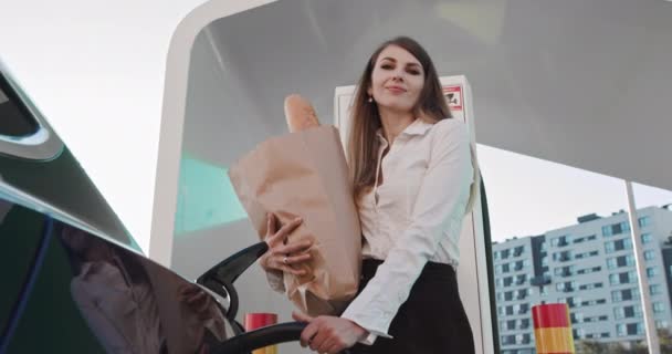 Una mujer se para con un paquete de productos cerca de su coche eléctrico y espera que el vehículo se cargue. Slow Motion La chica dirige su mirada a la cámara. Tecnologías Concepto Ambiental Renovable — Vídeos de Stock