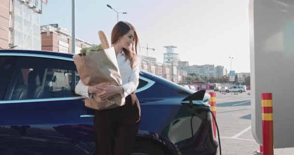 Красивая деловая женщина стоит рядом с ее электромобилем с продуктами. Зарядка электромобиля. Экологически чистый транспорт Завершение автоматического заряда. Высокоскоростная зарядная станция — стоковое видео