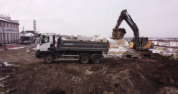高空空中射击:一个牵引桶充满卡车的身体.一台挖掘机和一辆卡车装载地面。飞越工人们倾泻地基的建筑工地.很多设备 — 图库照片
