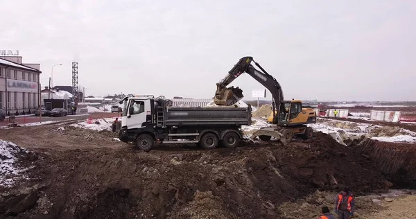 Övre antennen skott: en traktor hink fyller lastbilen kroppen. En grävmaskin och en lastbil lastar marken. Flyg över byggarbetsplatsen där arbetarna häller i grunden. Massor av utrustning — Stockfoto