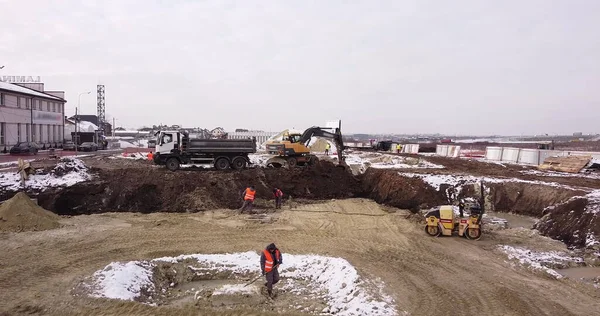 Верхній повітряний постріл: відро для трактора заповнює кузов вантажівки. Екскаватор і вантажівка завантажують землю. Політ над будівельним майданчиком, де робітники заливають фундамент. Багато обладнання — стокове фото