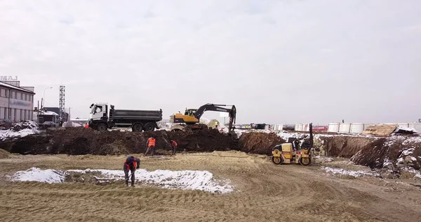 En stor byggarbetsplats i staden, den massiva byggnadsprocessen med tunga fordon i arbete, grävmaskin och bulldozer. Antenn ovanifrån: gul grävmaskin häller jord i tipper — Stockfoto