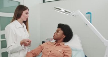 Kozmetik uzmanı hastayla iletişim kuruyor. Deri tedavisi ve bakımı. Hastane resepsiyonunda Kozmetologlar 'ın kürsüsünde oturan Afrikalı Amerikalı kadın hasta. Kızı mezoterapi prosedürüne hazırlıyorum.