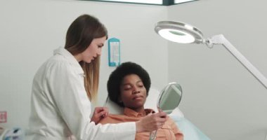 Güzellik kliniğinde aynaya bakan kadın müşteriye danışmanlık yapıyor. Güzellik endüstrisi konsepti. Güzellik uzmanı hastayı kaplıca salonunda cilt bakımına hazırlıyor. Kozmetoloji Ofisi