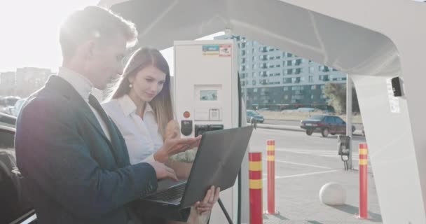 Mann und Frau arbeiten nach dem Einkaufen in der Nähe eines Elektroautos. Arbeiten Sie aus der Ferne, freiberuflich. Auto an der Ladestation. Erfolgreiche Menschen arbeiten auf dem Hof. Arbeit beim Laden des Autos. — Stockvideo